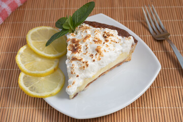 A slice of homemade lemon meringue pie on a plate, with slices of lemons, napkin completing the scene in a rustic setting and wooden planks background