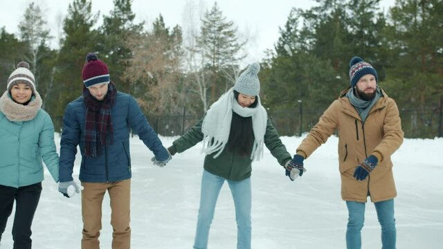 Girls and guys in warm clothes are ice-skating in park smiling holding hands having fun together. Green pine trees forest is visible in background.