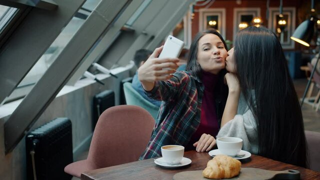 Charming young ladies are taking selfie with smartphone camera posing kissing having fun in cafe. Friendship and modern technology concept.