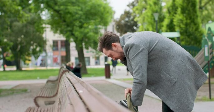 Young man with mustaches and a beard is searching for something in his backpack, placed on the wooden bench on the square. Green trees are on the background