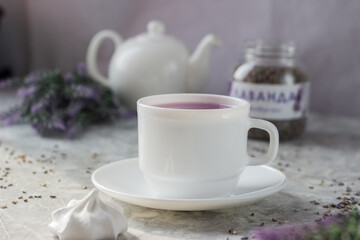 Fototapeta premium lavender tea in a white mug. Purple tea in a mug on a light background stands on the table next to lavender flowers. Dried lavender flowers are brewed in a Cup.