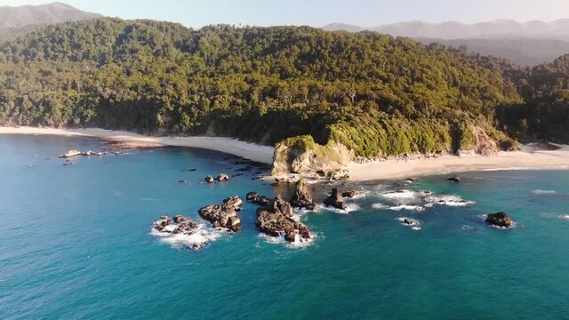 Aerial orbit reveal shot of New Zealand shore during golden hour. Landscape of jungle forest in contrast to white sandy beaches and rock formation. Monro Beach, West Coast