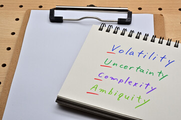 The words "Volatility, Uncertainty, Complexity, Ambiguity" are written in the notebook and underlined in the initials with the clipboard.