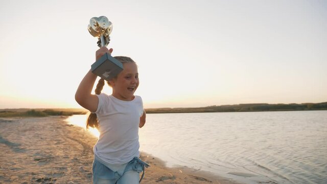 Happy kid winner runs along the beach at sunset with a cup in hand above his head. The kid celebrates the victory. The path to success is the highest happiness. Superhero child. Achievement award.