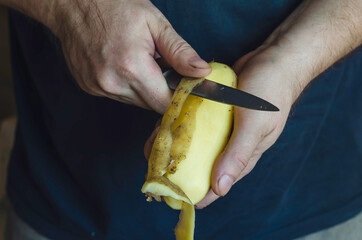 A mature man peels raw potatoes with a kitchen knife.