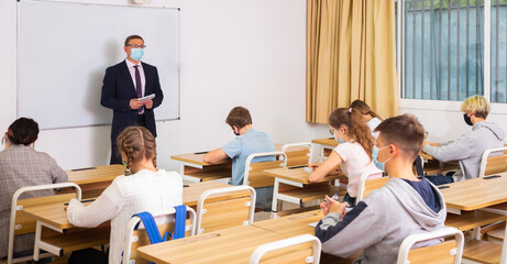 Focused male teacher in protective face mask giving lesson to teenage students in college. New life...