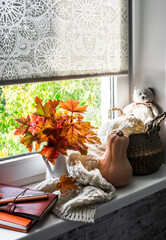 House window autumn still life - bouquet of maple leaves in a jug, straw basket with knitted blanket, pumpkin, books on the windowsill. Cozy interior design