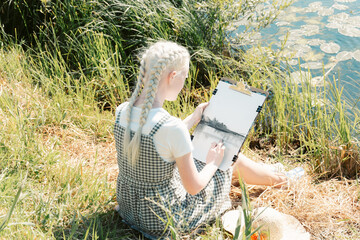 a young albino woman in a straw hat, sitting on the river Bank and draws a landscape on canvas with...