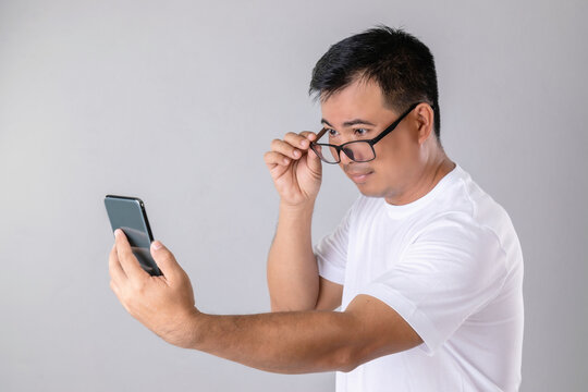 Short Or Long Sighted Concept : Man Weraing Eyeglasses And Trying To Look Clearly On Smarthphone In Studio Shot On Grey