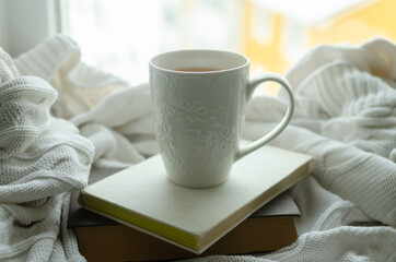 White cup of tea on the windowsill with books and white knit blanket