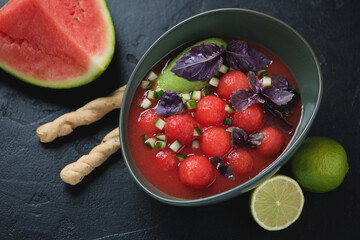 Bowl of watermelon and tomato gazpacho soup served with grissini, studio shot on a black stone background, elevated view