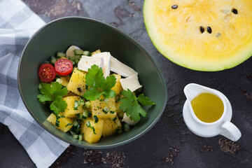 Green bowl with yellow watermelon, herbed cucumber and cheese salad, high angle view on a brown stone background, studio shot