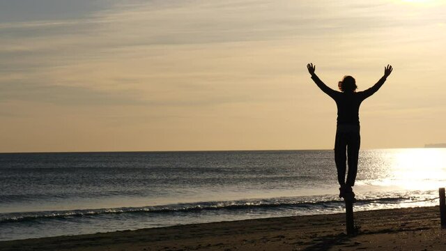 Silhouette of woman at dawn morning on beach, looking at sea horizon, enjoying sunrise with arms raised up. Back view.
