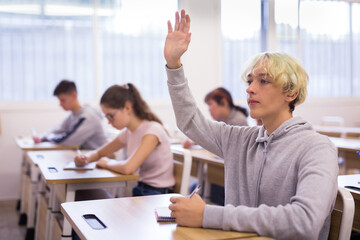 Naklejka na ściany i meble Portrait of smart teenage schoolboy raising hand to answer during lesson in college