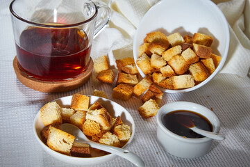 Square toasted pieces of homemade delicious rusk, hardtack, Dryasdust, zwieback, Liquid honey in a saucer and black tee in a cap on a white tablecloth.