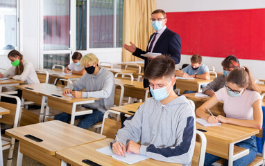 Focused teenage students in protective face masks studying in classroom with teacher, writing...