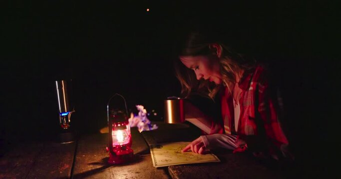 Young woman in red classic shirt sitting at wooden table by campfire looking on the map and drinking hot tea enjoying outdoor adventure. Red lantern lightning the night scene. Woman on camping