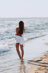 Young beautiful slender woman in a black bikini and white shirt on a tanned body on the beach, back view. Soft selective focus.