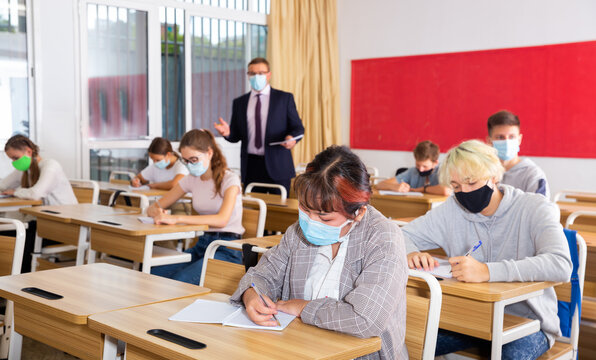 Teacher In Protective Mask Explains To The Students The Subject Of Instruction In The Classroom