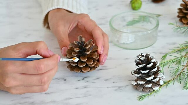 Christmas Crafts. Female hands decorate pine cones with white paint. Close-up.