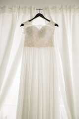 Long white dress of the bride with a lace corset on a black hanger against a background of white curtains.