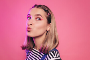 Positive amazing young caucasian woman blowing a kiss to someone and looking away. Isolated over pink background. Lips in a tubule. Bright makeup, smart casual style concept.