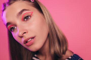 Fototapeta premium Attractive young woman with tired expression on her face bowed her head to one side. Close-up view of pretty millenial girl with light brown shloulder-length hair and nose piercing. Pink background.