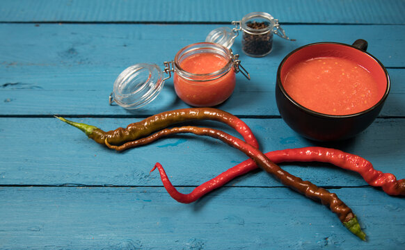 Red Hot Pepper Pods, Red Pepper Sauce In A Glass Jar And Cup On A Wooden Blue Background. Place For A Copy Space