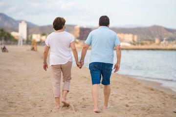 pensioner woman and her husband taking romantic walk together - happy retired mature couple in face mask walking on the beach in new normal holidays trip during covid19