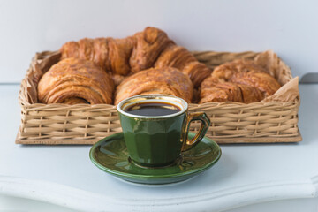 Traditional french breakfast. Vintage green cup of coffee and basket with hot croissants on white table
