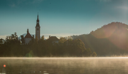 Low profile panoramic photo of lake Bled island with church on a cold hazy foggy early autumn morning. Visible fog coming out of water.