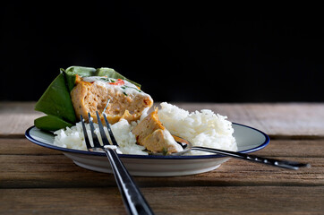 Cooked rice and Steamed fish with curry paste in banana leaf cup and *Yor leaves (Thai herb)* in white dish on old wooden table which has black background