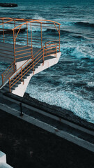 A white pier with yellow railings overhanging the swirling blue sea. Big foam waves. Vertical photo.