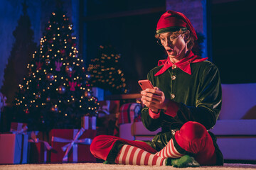 Full size photo of amazed elf sit floor read x-mas news on cellphone in house indoors with...