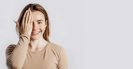 Young beautiful girl smiles and covers half of her face with her hand on a white isolated background. A woman points to an idea, a place for advertising. Positive brunette in a beige jumper.