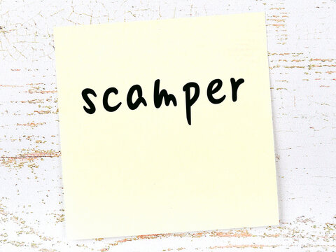 Yellow sticky note on wooden wall with handwritten word scamper