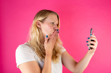 Blonde young woman holding smartphone while making a video call on pink background. 