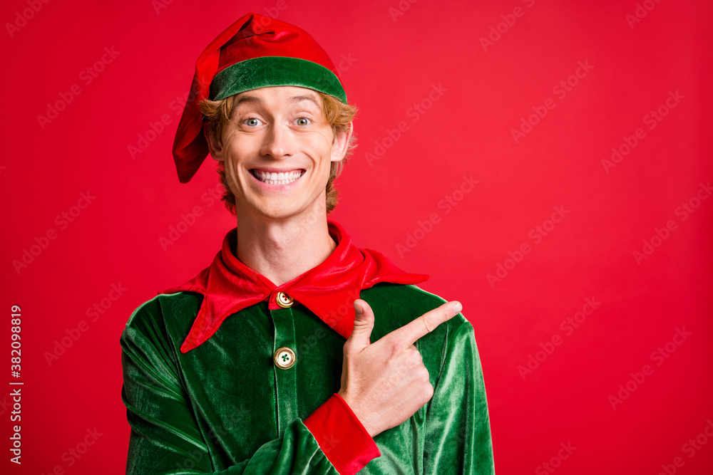 Wall mural Photo of positive elf guy point finger empty space x-mas christmas ads wear costume cap isolated on bright red color background