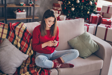 Obraz premium Full length photo of positive girl sit couch text cellphone wear red sweater in house indoors with x-mas christmas decoration