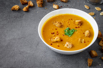 Pumpkin soup with coriander, gluten free croutons, chili and garlic flakes seasoning in a white bowl. Croutons and pumpkin seeds in the background. Seasonal autumn / winter food. Space for text