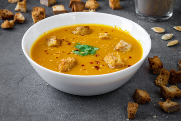 Side view of pumpkin soup with coriander, gluten free croutons, chili and garlic flakes seasoning in a white bowl. Croutons, pumpkin seeds and a candle in the background. Autumn / winter food