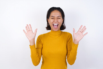 Emotive  Young hispanic girl with short hair wearing casual yellow sweater isolated over white background laughs loudly, hears funny joke or story, raises palms with satisfaction, being overjoyed