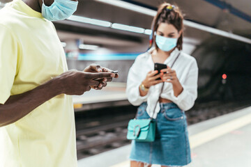 Close-up of black man with protective mask on face using smart phone while standing at subway station against woman