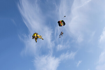 Skydiving. There are two skydivers in the sky. First skydiver is deploying a parachute, second skydiver is speed flying in the blue sky.