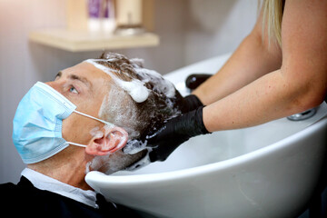 Female hairdresser hands in rubber gloves washes shampoo the head of a medical mask man.