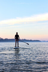 Back view on teenager boy rowing on SUP (stand up paddle board) in autumn Danube river at foggy morning against the backdrop of autumn trees without leaves