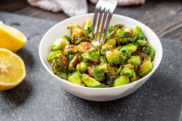 Woman or man eating roasted / fried brussels sprouts from a white bowl with chili and garlic flakes seasoning using a fork. Lemon halves and towel sitting on a cutting board in the background.