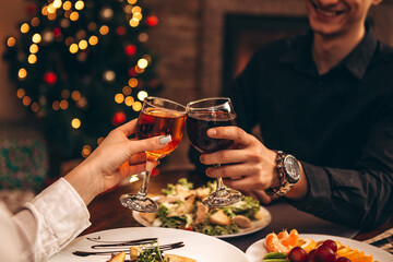 hands with glasses clinking against the background of Christmas tree lights and bonfires from a home fireplace over a table with delicious dishes