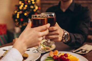hands with glasses clinking against the background of Christmas tree lights and bonfires from a home fireplace over a table with delicious dishes
