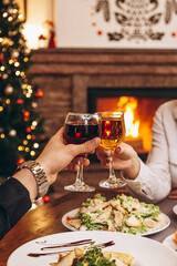 close up hands with glasses clinking against the background of Christmas tree lights and bonfires from a home fireplace over a table with delicious dishes